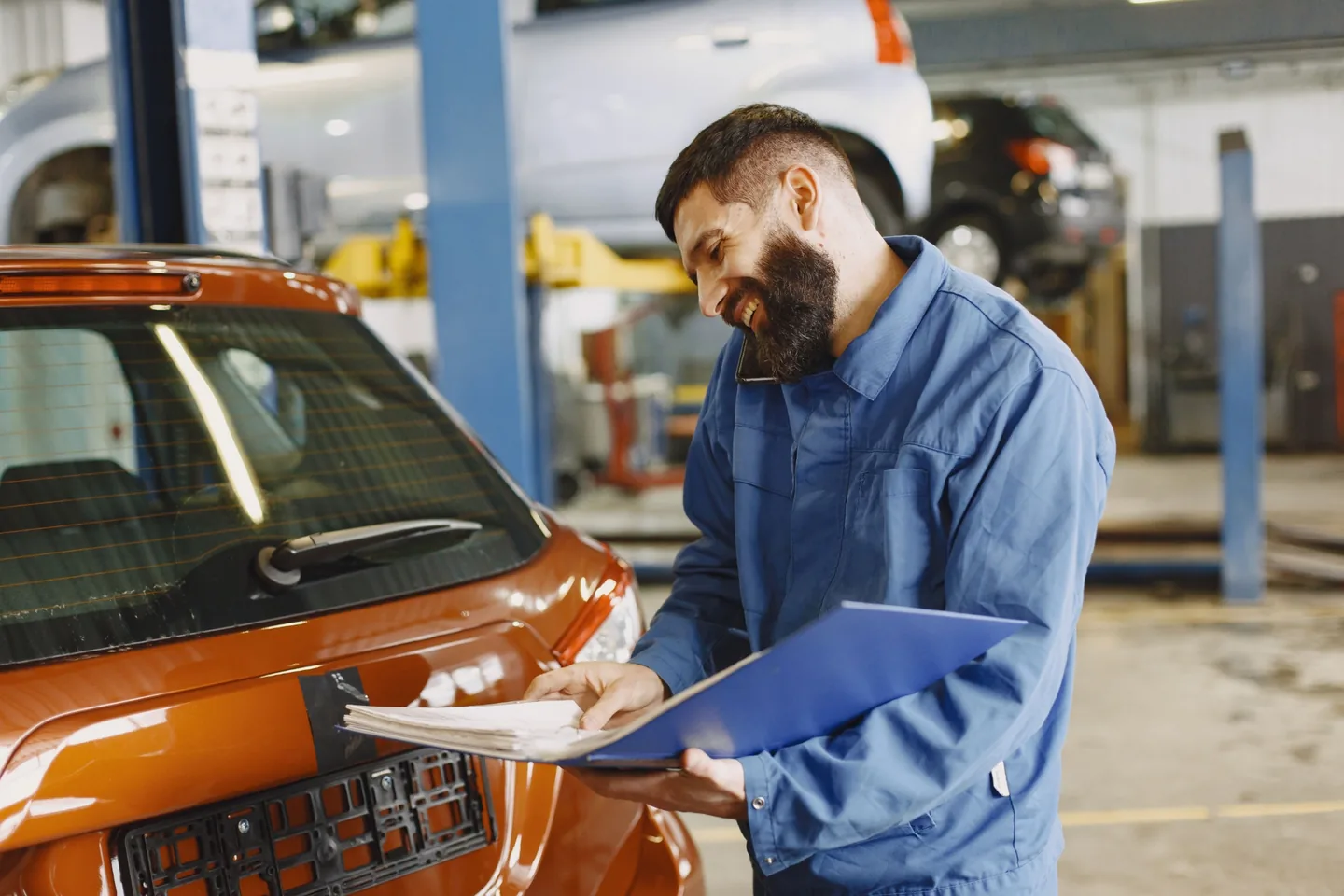 An automotive technician talking on the phone while reviewing paperwork beside a car
